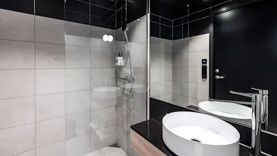 Ett modernt badrum med en elegant vit vask på en mörk bänkskiva speglas i en stor spegel. En dusch med glasväggar och silverfästen finns mot grå kakelväggar.