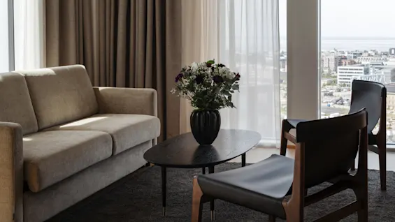 Et koselig oppholdsrom med sofa, salongbord med blomster og stoler, med panoramautsikt over byen fra et stort vindu.