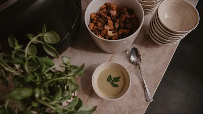 En liten skål suppe med en garnityr står på en kjøkkenbenk ved siden av en skje, krutonger, stablede tomme skåler og fersk basilikum.