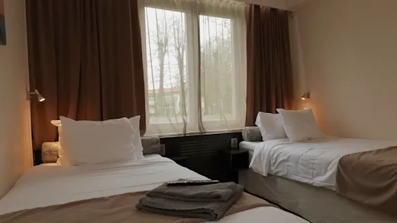 To pænt redte enkeltsenge i et hotelværelse, dækket med hvide lagner og brune tæpper, vender mod et vindue med brune gardiner. En fjernbetjening ligger på en seng.