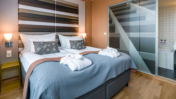 A plush bed is neatly arranged with pillows and folded robes, set against a decorative panel in a modern hotel room, adjacent to a translucent glass partition leading to a tiled bathroom.