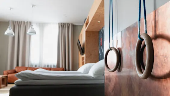 Gymnastics rings hang from a wall in a modern bedroom, featuring a neatly made bed, a brown sofa, pendant lights, and large curtains.