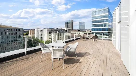 Rooftop terrace with round tables and chairs, set against a cityscape of modern glass buildings and clear blue skies.