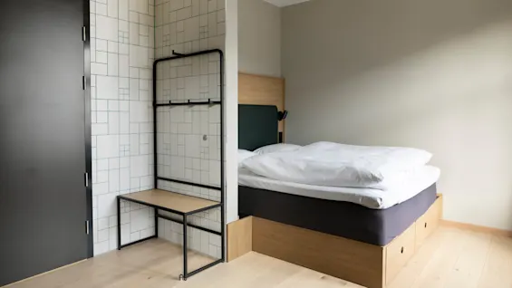 En säng med vita lakan står i ett minimalistiskt rum, bredvid en mönstrad väggpanel och en svart dörr, vilket skapar en modern och luftig atmosfär.