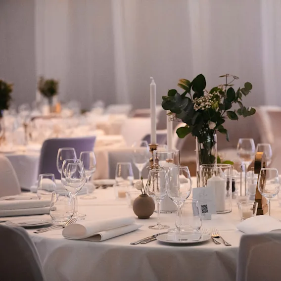 Elegant bryllupsbord dekket med hvit duk, høye levende lys, vinglass og grønne eukalyptusdekorasjoner.