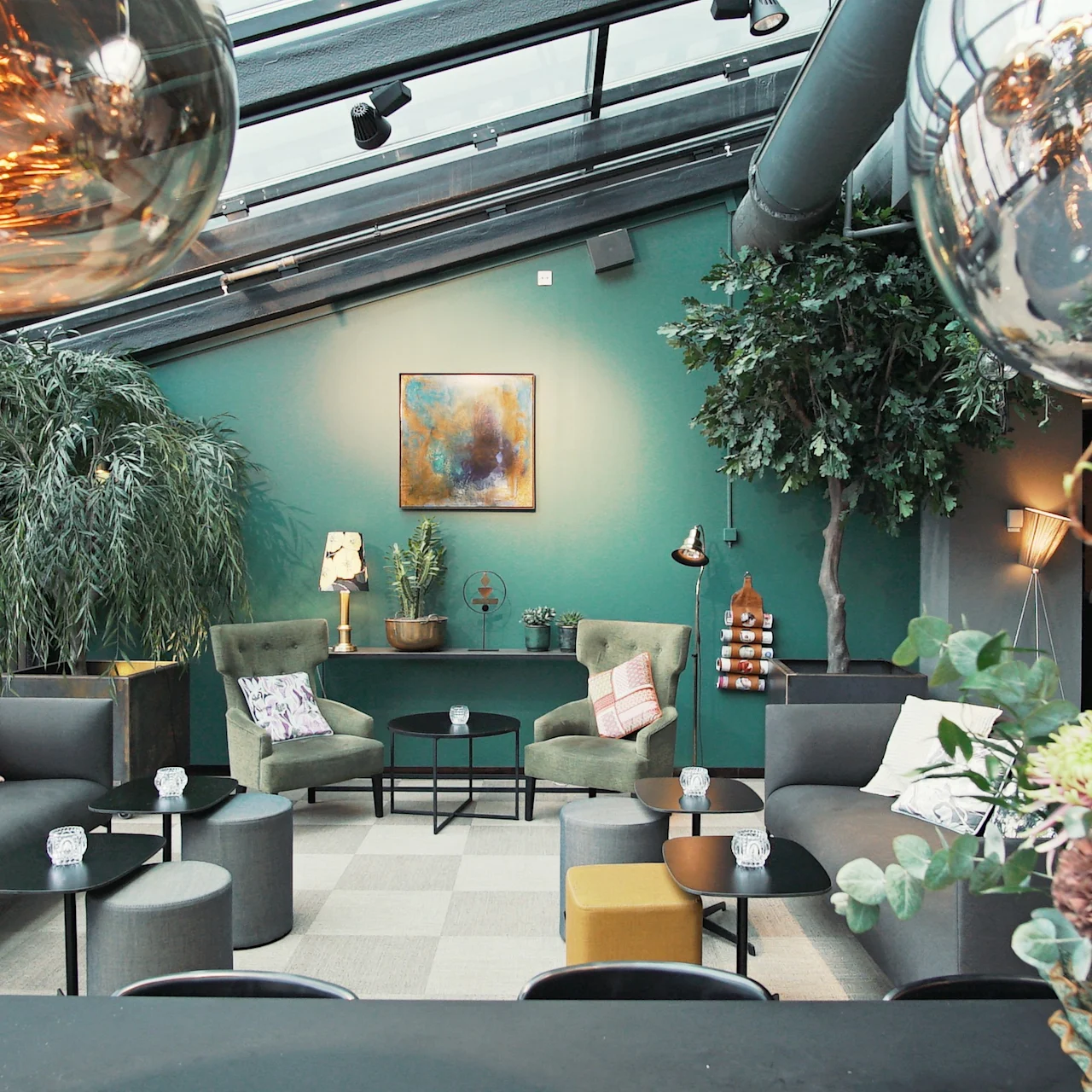 Lyxig lounge med moderna soffor i ett rymligt rum med växter och kupolformade taklampor. Konst pryder turkosa väggar och skapar en mysig, inbjudande atmosfär.