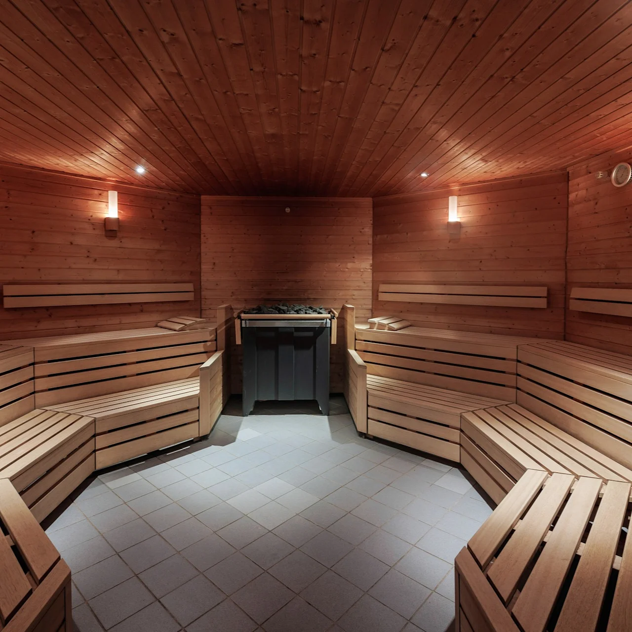 A wooden sauna with rows of benches wraps around three sides, surrounding a central heater with stones, illuminated by soft wall lights, creating a warm and inviting atmosphere.