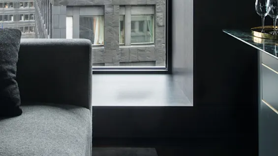 En grå sofa står ved siden av et stort vindu som vender ut mot en moderne grå bygning. Et glassbord med vinglass er i nærheten.