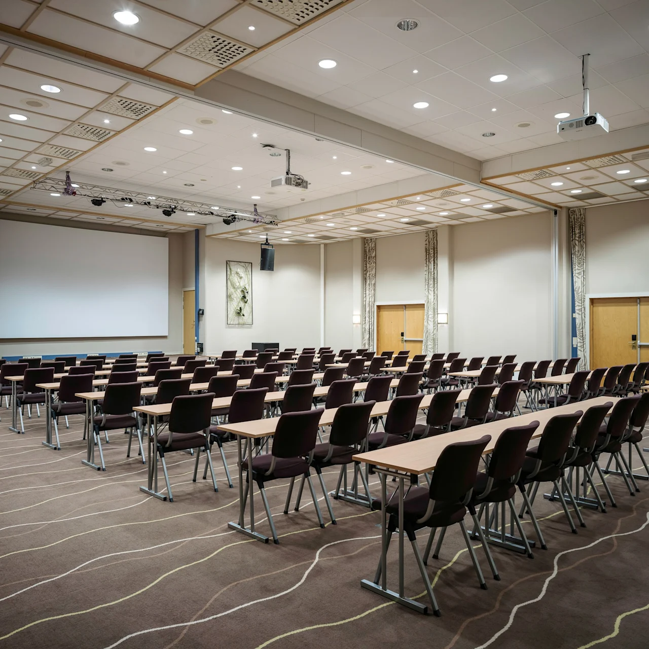 Rows of chairs and tables face a large projection screen in a conference room with carpeted flooring, overhead lighting, and decorative wall panels.