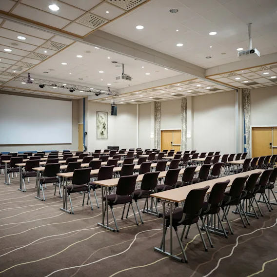 Rader av stoler og bord vender mot en stor projeksjonsskjerm i et konferanserom med teppebelagt gulv, overbelysning og dekorative veggpaneler.