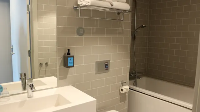 Handfat med spegel och tvålpump i modernt badrum; handdukar är prydligt vikta på en hylla, badkar med duschhuvud i bakgrunden, gråa tunnelbanekakel på väggarna.