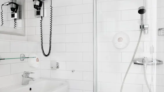 En hvit keramisk vask med kromkran er montert på en flisbelagt baderomsvegg, ved siden av en glasshylle og en dusj med håndholdt hode.