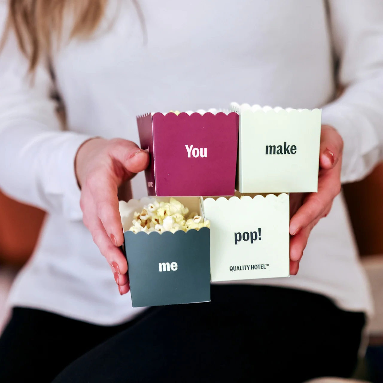 Boxes labeled "You make me pop! QUALITY HOTEL™" are held by a person in a white shirt, containing popcorn, against a casual indoor backdrop.