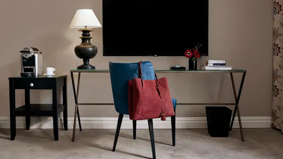 En blå stol med en rød taske står ved et glassbord med en lampe, røde blomster og bøker. I nærheten står et lite bord med en kaffemaskin og kopp under en veggmontert TV.