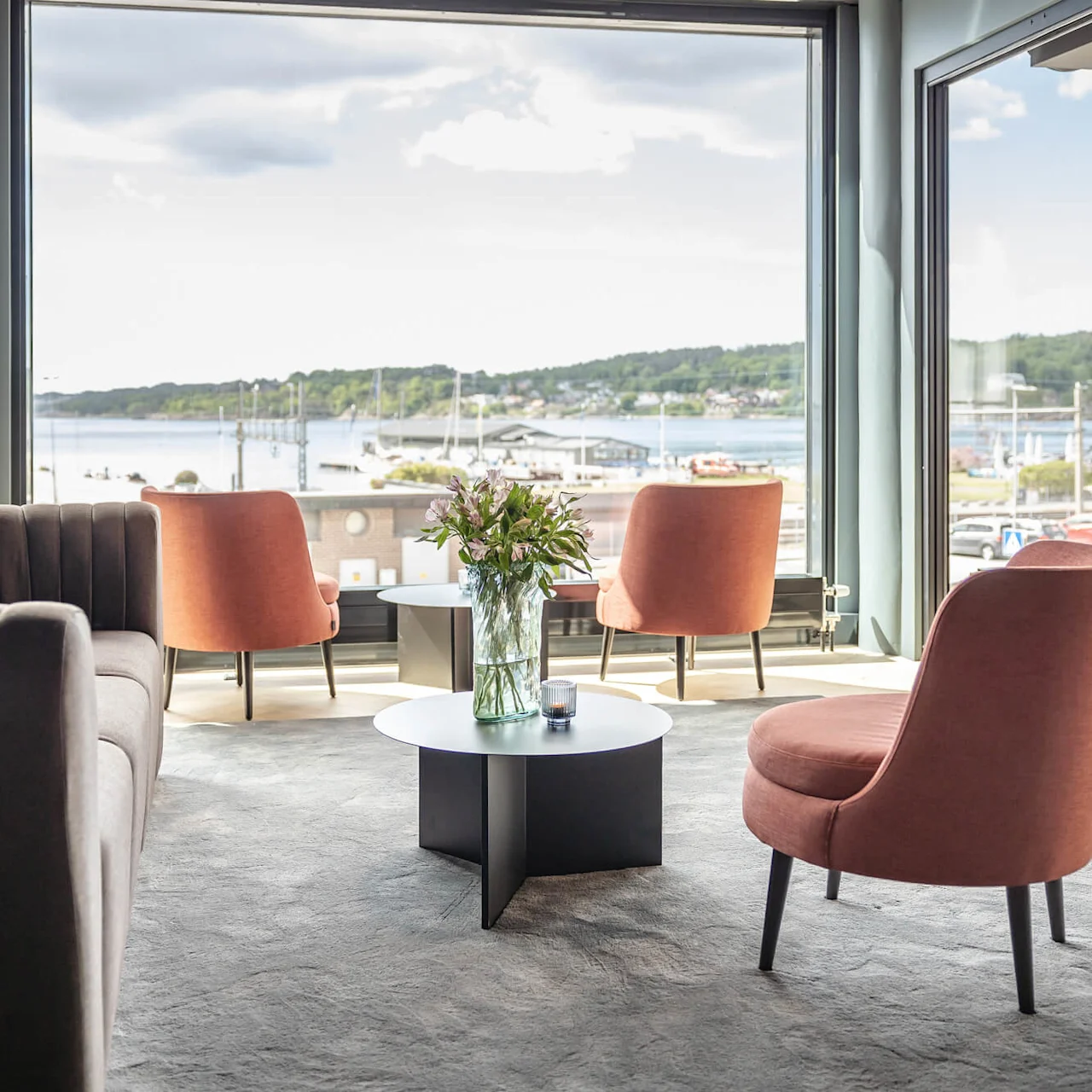 Sofistikeret siddeområde med moderne stole og en sofa omkring et bord med blomster. Store vinduer giver en naturskøn udsigt over vandet under en delvist overskyet himmel.