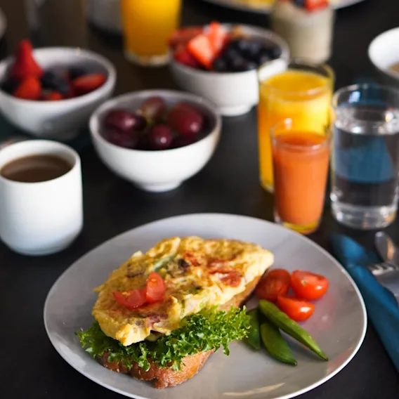 Omelet on toast garnished with lettuce and cherry tomatoes sits on a plate. Surrounding are bowls of fruit, cups of coffee, and glasses of orange juice on a dark table.