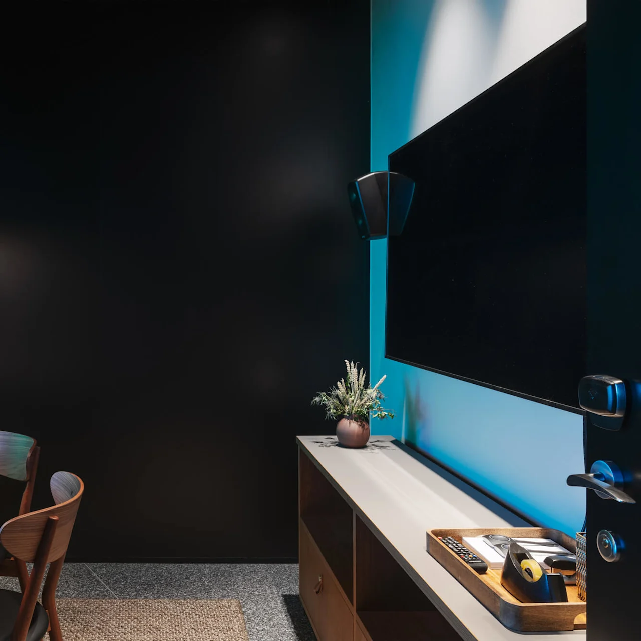 A round table with wooden chairs sits in a dimly lit room. A television is mounted on a wall beside a cabinet with decor and office supplies.