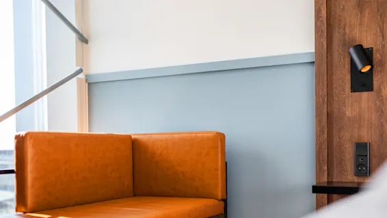 En brun skinnbenk hviler mot en lyseblå vegg, ved siden av en treplate med en liten svart lampe og en stikkontakt. Sollys filtreres gjennom et nærliggende vindu.