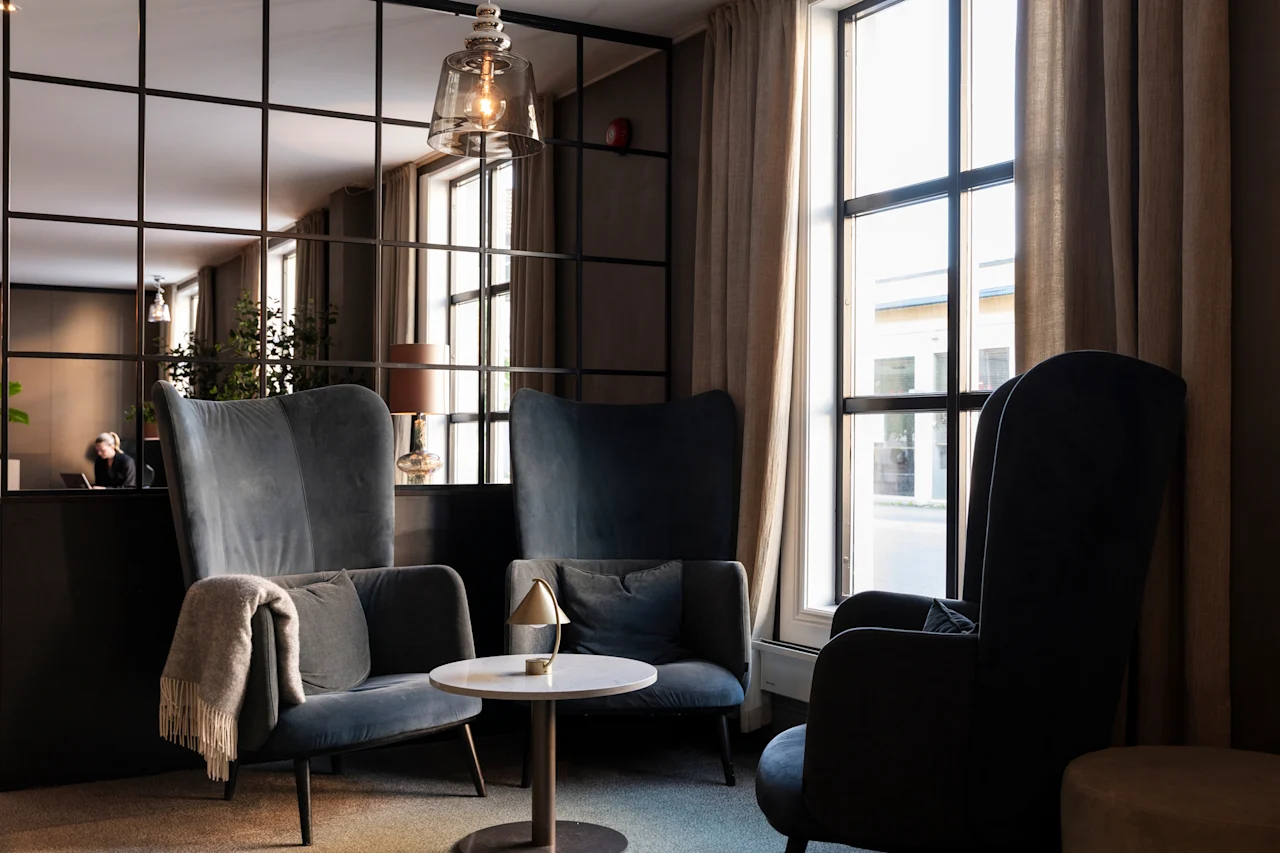 A comfortable lounge space featuring plush armchairs, a small table, and a bright window, with a person working nearby.