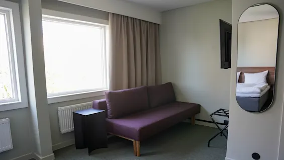 En lilla sofa står mot en lys vegg ved siden av et svart sidebord i et rom med et speil, vindu og en delvis synlig seng.