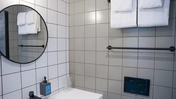 Et rundt speil henger på en flislagt baderomsvegg over en vask med sort kran og såpedispenser merket "HAND WASH". Håndklær er pent brettet på en stang.