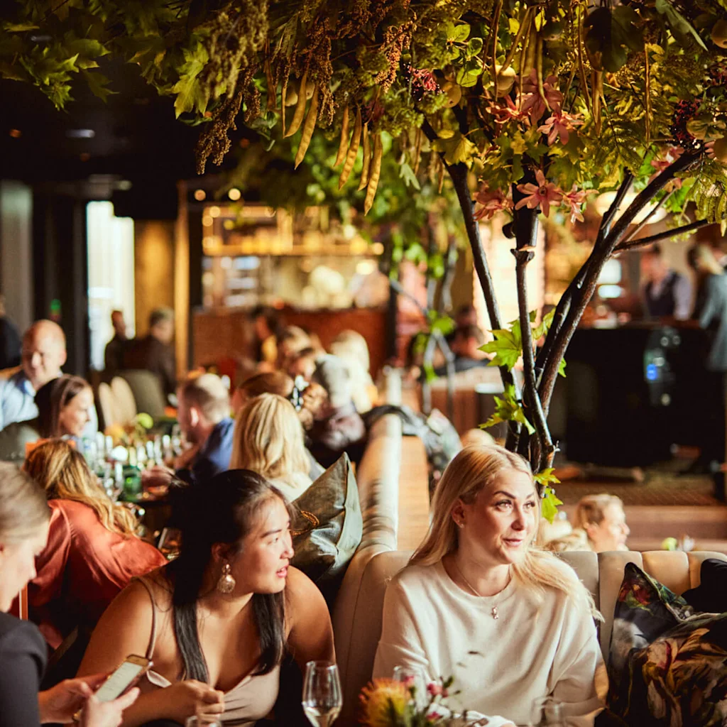People enjoying a meal and conversation in a lively restaurant with lush green and flowering decorations overhead.