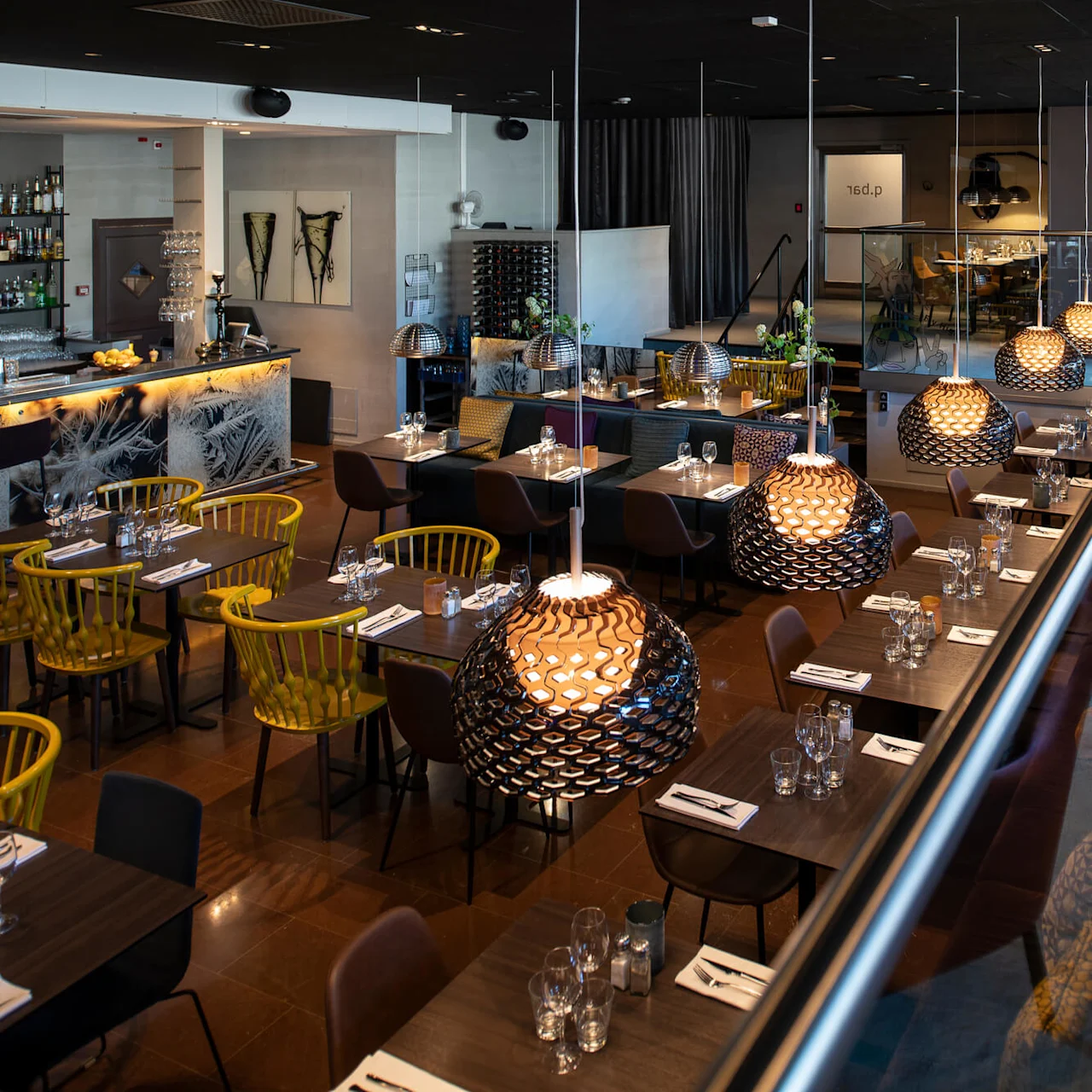 En modern restaurang med dukade bord och stilfulla hängande lampor. Barområde synligt till vänster, färgglada stolar ger liv. Glasvägg skiljer sittplatserna. 