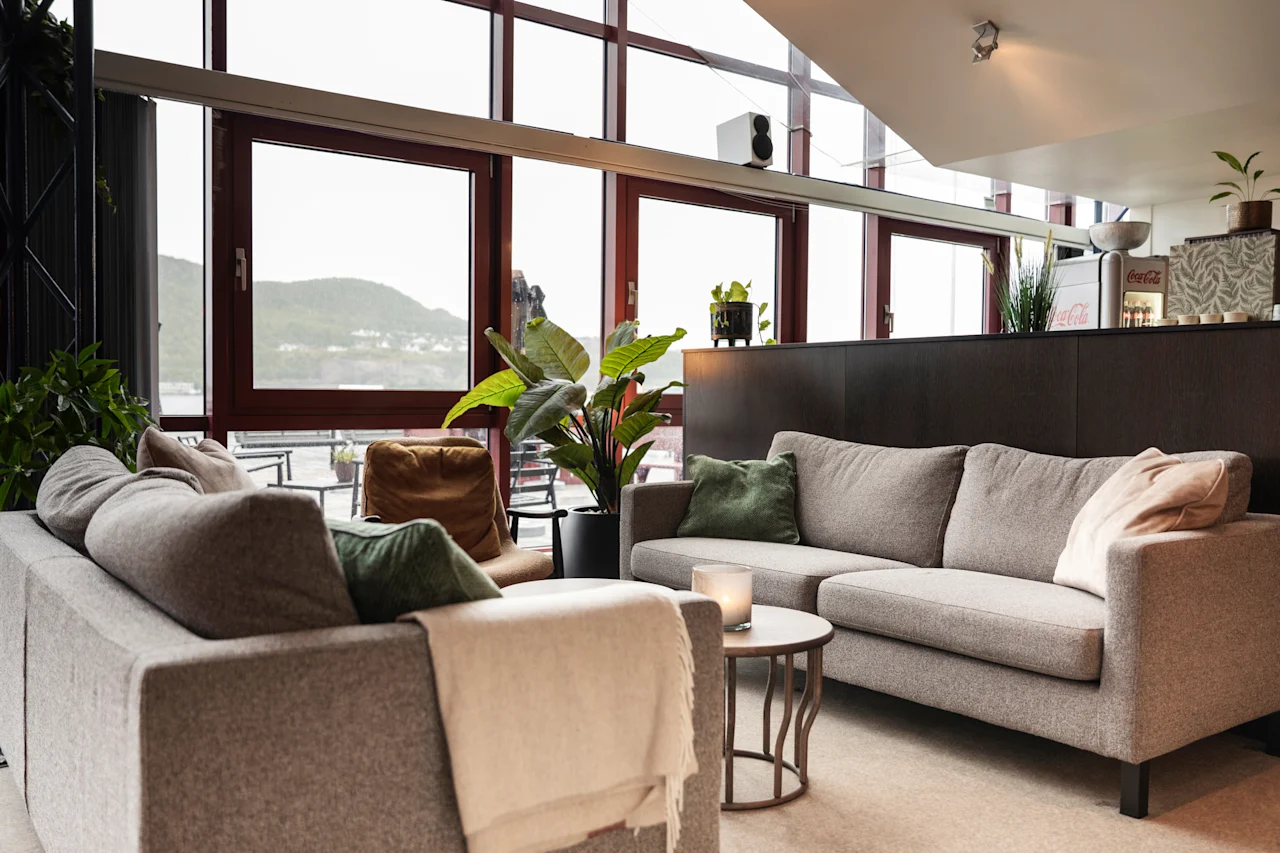 Mukava oleskelutila, jossa on muhkeat sohvat, viherkasveja ja suuret ikkunat, joista avautuu näkymä vuorille ja vedelle. Viihtyisä ja kutsuva tila.