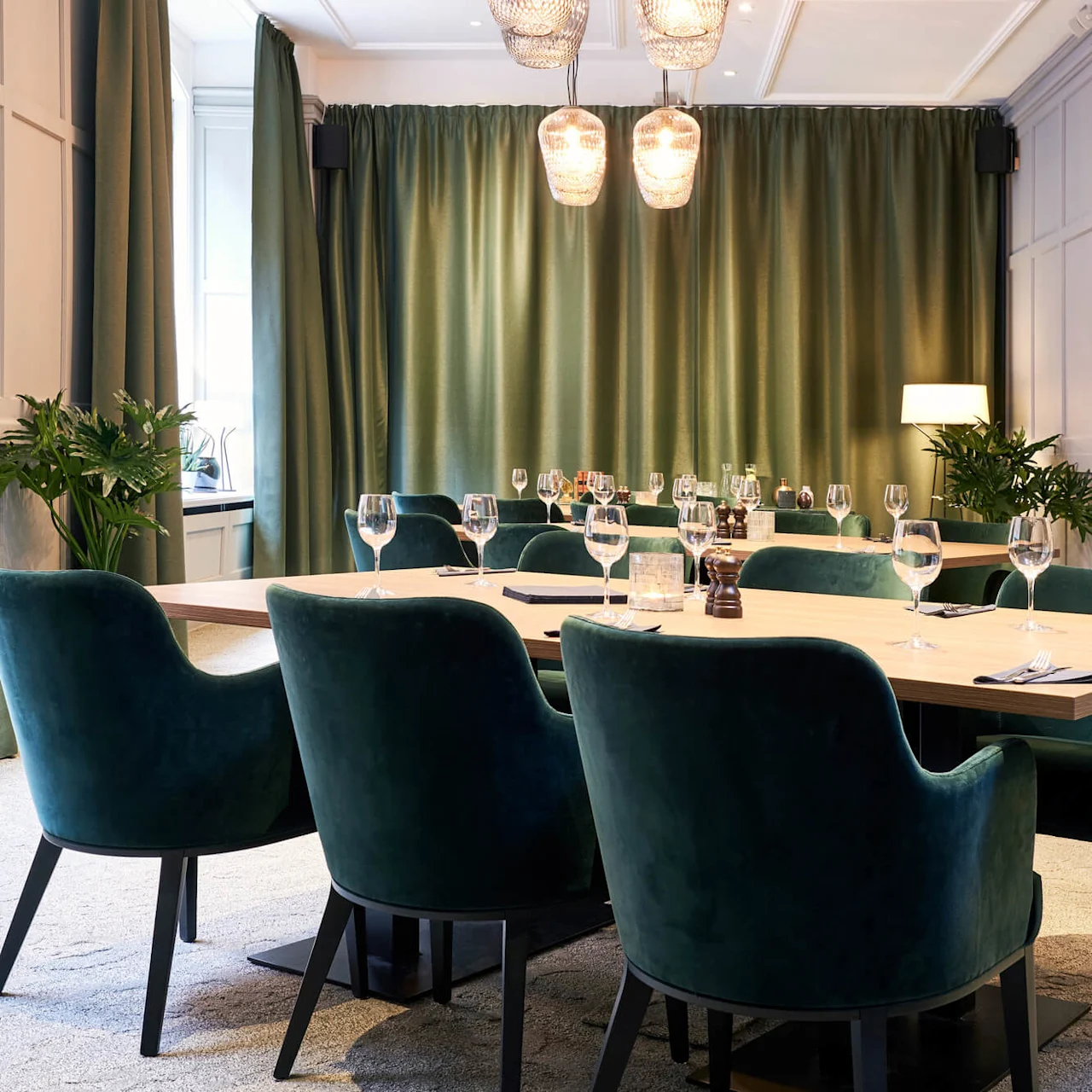An elegantly set dining table with green velvet chairs in a room with green curtains and panelled walls.