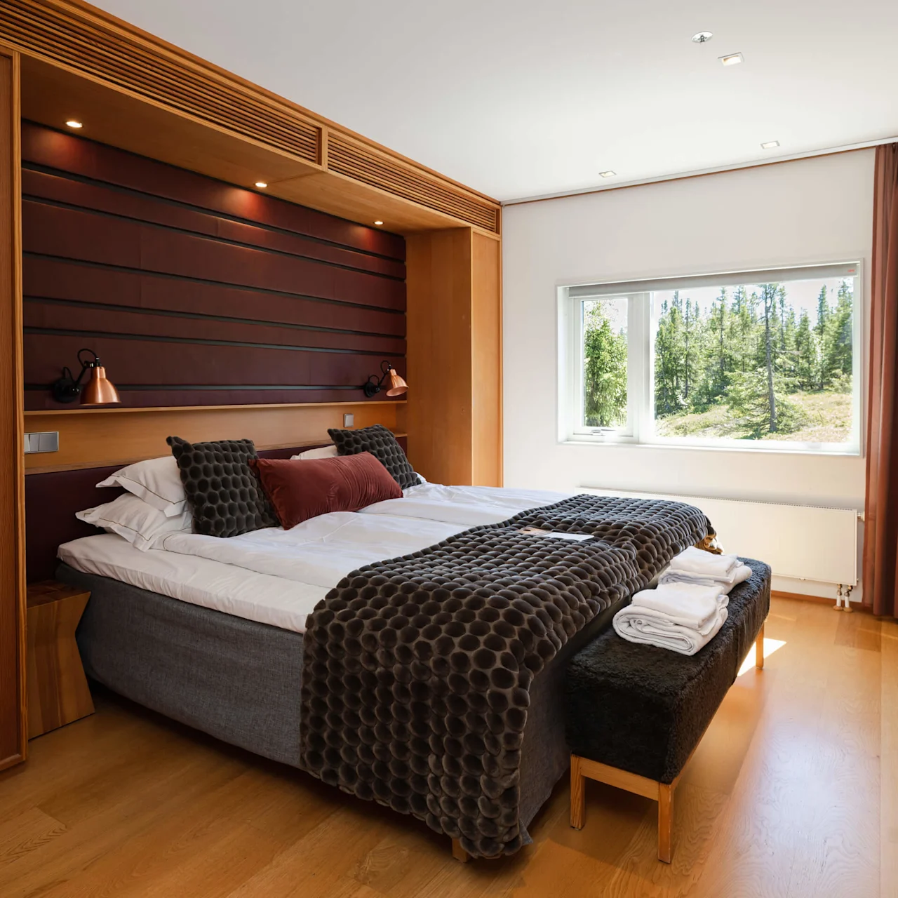 A neatly made bed in a bright, wood-paneled bedroom with a window showing a forest view.