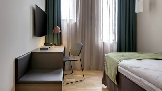 A bed with green bedding sits beside a window with sheer curtains, a wall-mounted TV, and a desk with a chair and lamp in a minimally decorated room.