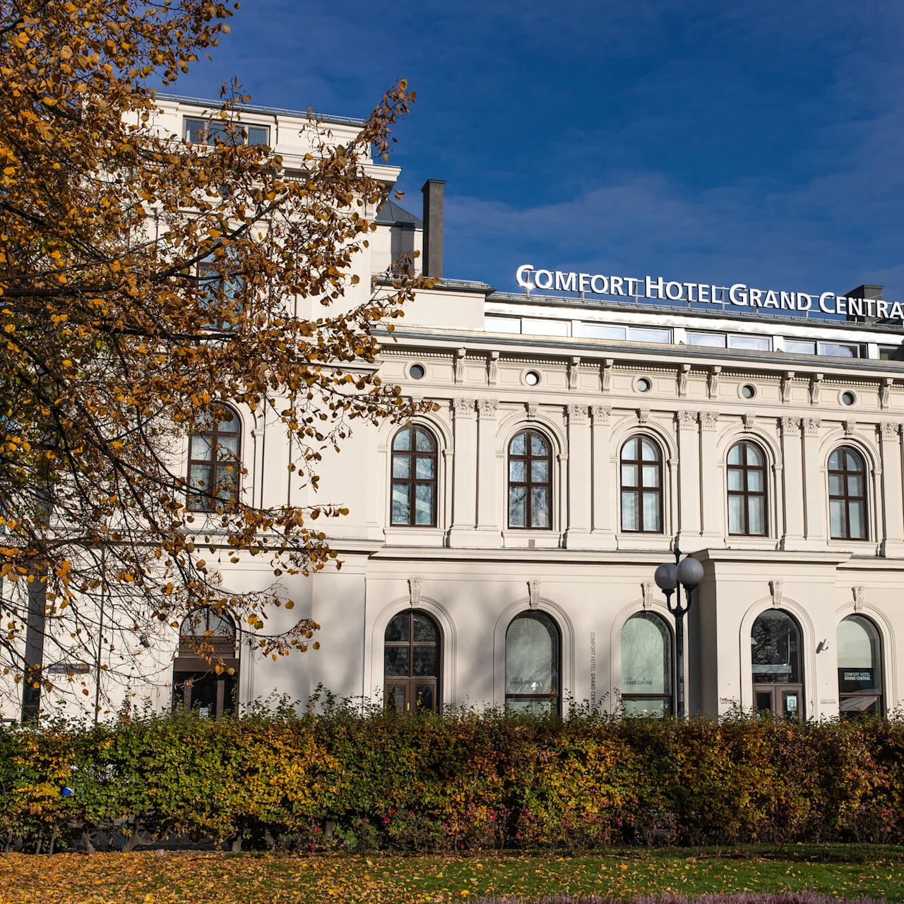 A grand hotel building stands surrounded by autumn trees and bushes, under a clear blue sky. Sign reads: "COMFORT HOTEL GRAND CENTRAL."