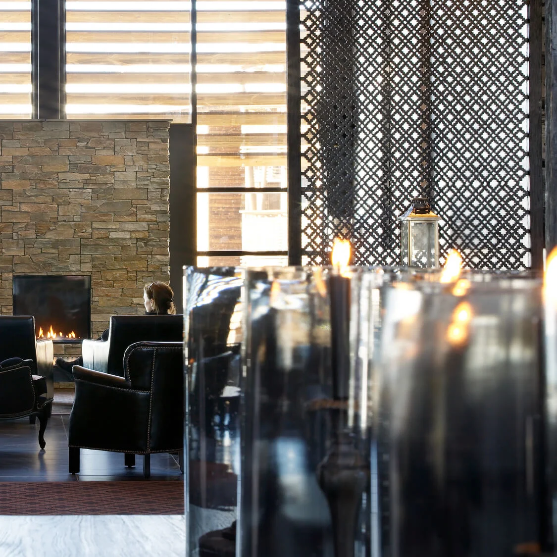 A cozy lounge scene shows a person sitting in a black armchair facing a stone fireplace with lit candles. The area is surrounded by patterned walls and large windows.