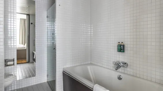 A modern bathroom with white tiled walls and floor, featuring a bathtub, toilet, and a glass shower partition. A mirror reflects a bedroom.