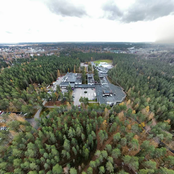 Suuri rakennuskompleksi on tiheän metsän ympäröimänä pilvisen taivaan alla, ja edustalla näkyy tie. Näkymä laajenee laajoihin metsämaisemiin.