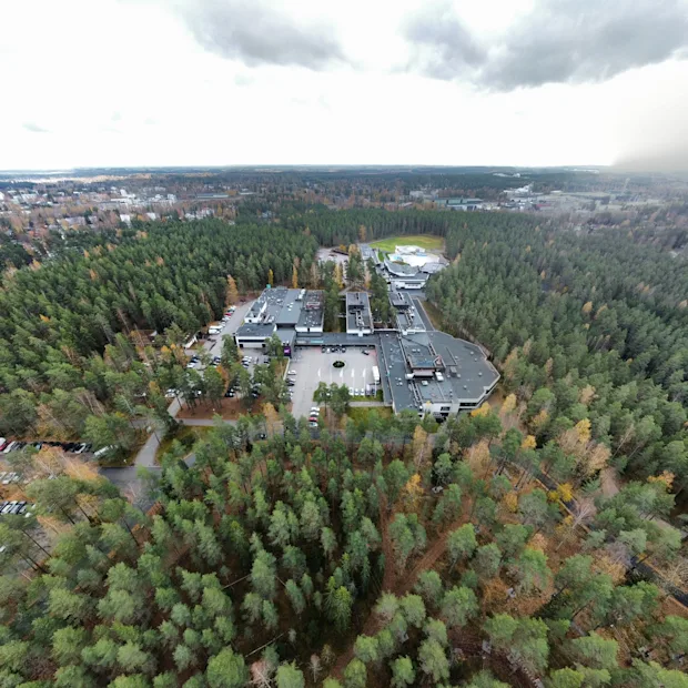 Suuri rakennuskompleksi on tiheän metsän ympäröimänä pilvisen taivaan alla, ja edustalla näkyy tie. Näkymä laajenee laajoihin metsämaisemiin.