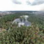 Suuri rakennuskompleksi on tiheän metsän ympäröimänä pilvisen taivaan alla, ja edustalla näkyy tie. Näkymä laajenee laajoihin metsämaisemiin.