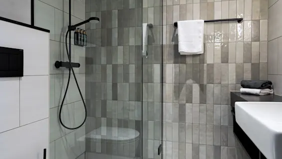 A modern bathroom features a glass shower with black fixtures. Towels rest on a rack and a sink is adjacent. Beige tiles cover the walls, providing a sleek, minimalist look.