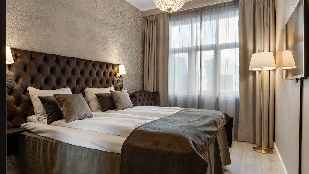 Et komfortabelt og elegant hotelværelse med en stor seng, tuftet sengegavl, lysekrone og vindue med gardiner.