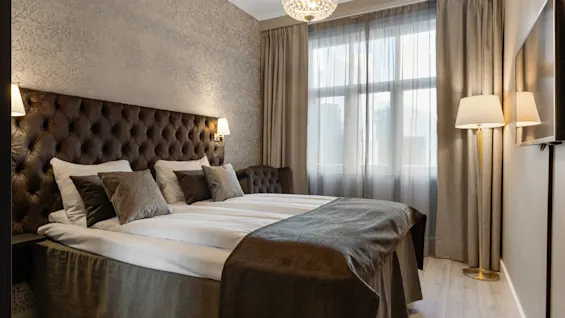 Et komfortabelt og elegant hotelværelse med en stor seng, tuftet sengegavl, lysekrone og vindue med gardiner.