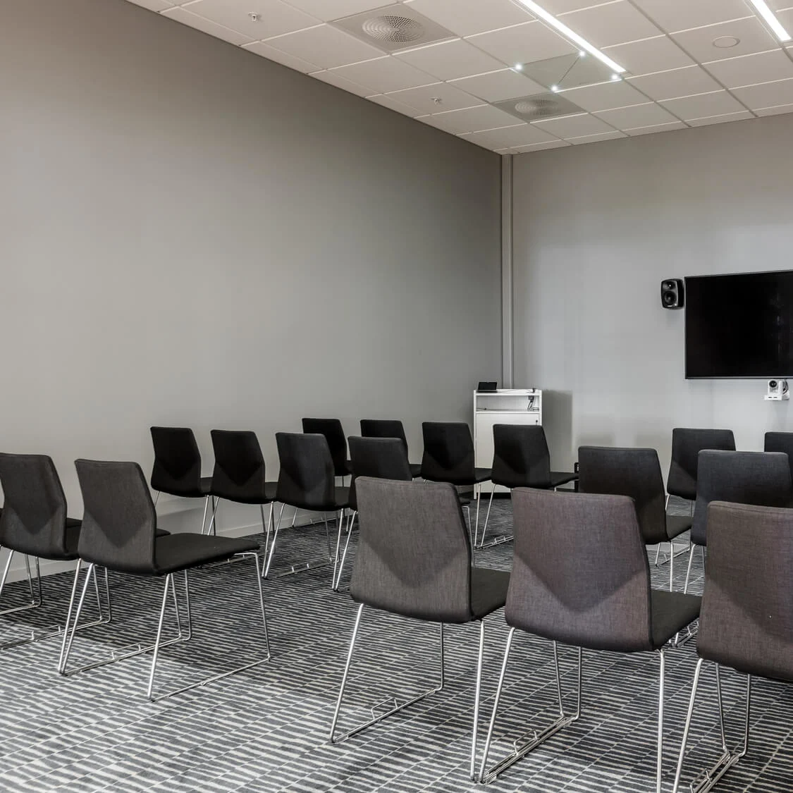 Rækker af sorte stole vender mod en vægmonteret TV med højtalere i et konferencelokale med grå vægge. En podium står i nærheden, og der er et tøjstativ i hjørnet.