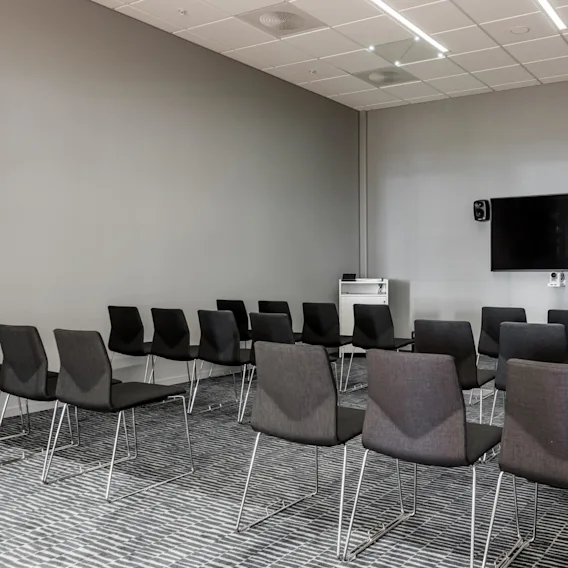 Rows of black chairs face a wall-mounted TV with speakers in a gray-walled conference room. A podium stands nearby, and a coatrack is in the corner.
