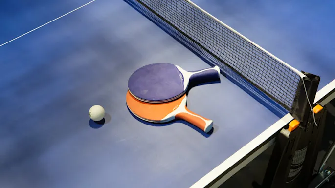 Ping pong paddles rest on a blue table, accompanied by a ball near the net.