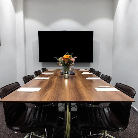 Et konferencebord af træ er dækket med papir og stole i et moderne mødelokale. En stor skærm er monteret på væggen, omgivet af minimalistisk indretning.
