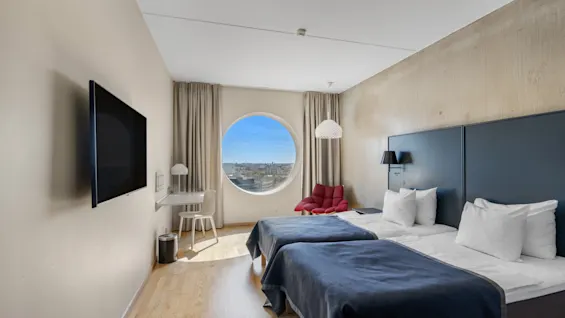 Et moderne hotelværelse badet i naturligt lys, med to enkeltsenge, et rundt vindue med byudsigt og et skrivebordsområde.
