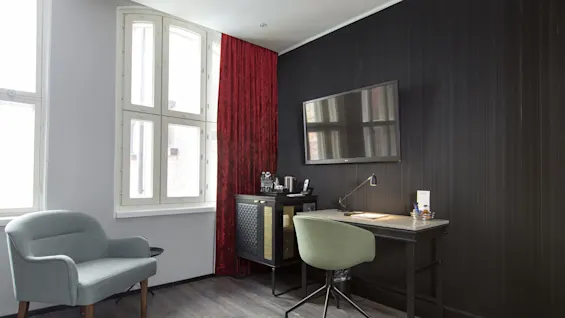 A furnished room with an armchair, a desk with a chair, a flat-screen TV, and a red curtain beside a window.