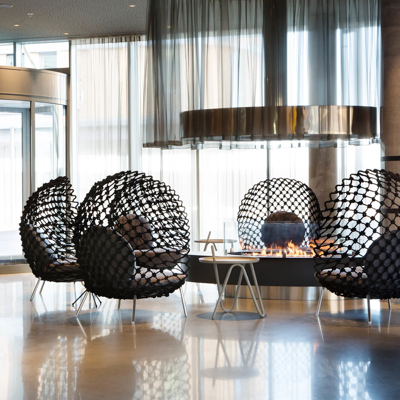 Woven black chairs encircle a modern fireplace, with flames flickering, in a sleek, minimalist interior featuring transparent curtains and a glass entrance.