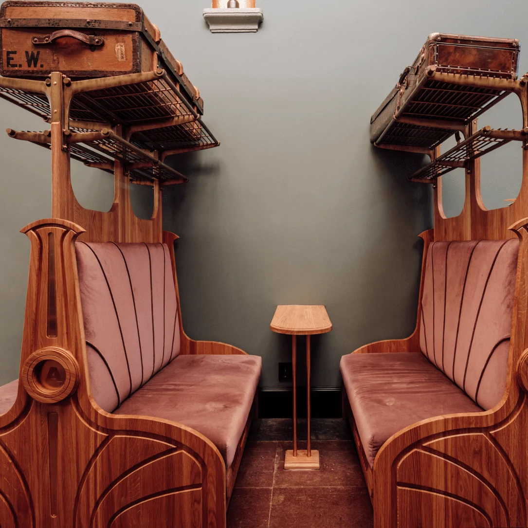 Two wooden benches with purple cushions face each other in a small room. Overhead luggage racks hold vintage suitcases. A small wooden table stands between the benches.