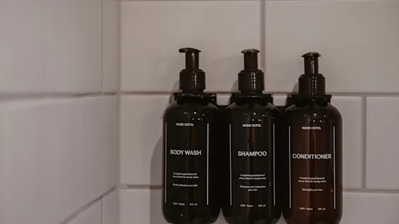Kolme tummanruskeaa pumppupulloa, joissa lukee Body Wash, Shampoo ja Conditioner, kiinnitettynä valkoiseen kaakeloituun suihkuseinään.