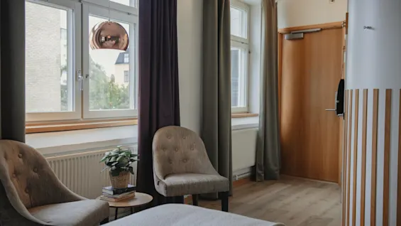 A cozy hotel room at Home Hotel Uppsala with a bed, two chairs, a plant, and large windows with curtains.